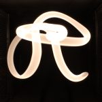 In a dark room, a streak of light forms a swirling script letter R to either side a chain link fence is lightly illuminated by the glow.