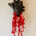 White background. In the middle, a bronze anatomical human heart hanging on a white wall. Red fiber yarn is crocheted and coming out of lower portions of the heart, hanging down. The red yarn has the appearance of blood dripping.