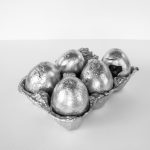 White background. Silver (aluminum) half dozen egg carton and five eggs. The carton and the eggs look broken and damaged.
