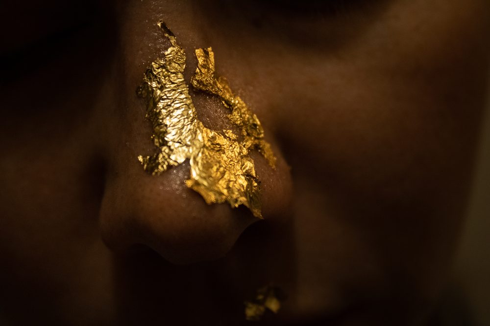 Macro photo of a nose covered in gold leaf flakes. The background is dark and the identity of the person is obstructed.