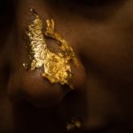 Macro photo of a nose covered in gold leaf flakes. The background is dark and the identity of the person is obstructed.