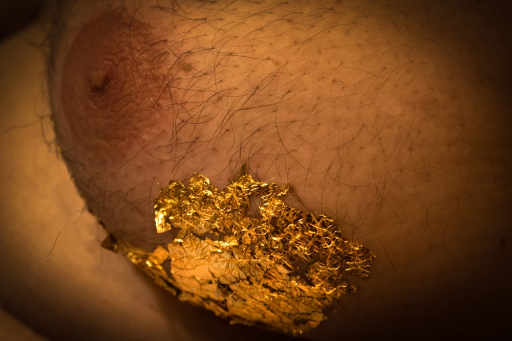 Macro photo of one side of a man's chest, profile view of the nipple, areola, and chest hair, with gold leaf flakes on the underside of the man's breast tissue.
