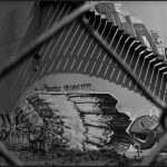 A photograph of a fence, and hidden behind the fence is a structure filled with graffiti.