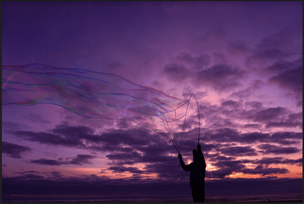 A photograph of a stranger blowing bubbles on the ocean front, capturing color from the sunset, and the sparkles shining from the bubbles.