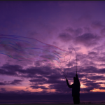 A photograph of a stranger blowing bubbles on the ocean front, capturing color from the sunset, and the sparkles shining from the bubbles.