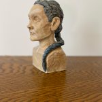 Ceramic representation of a middle aged person with a blank gaze and a blue braid.