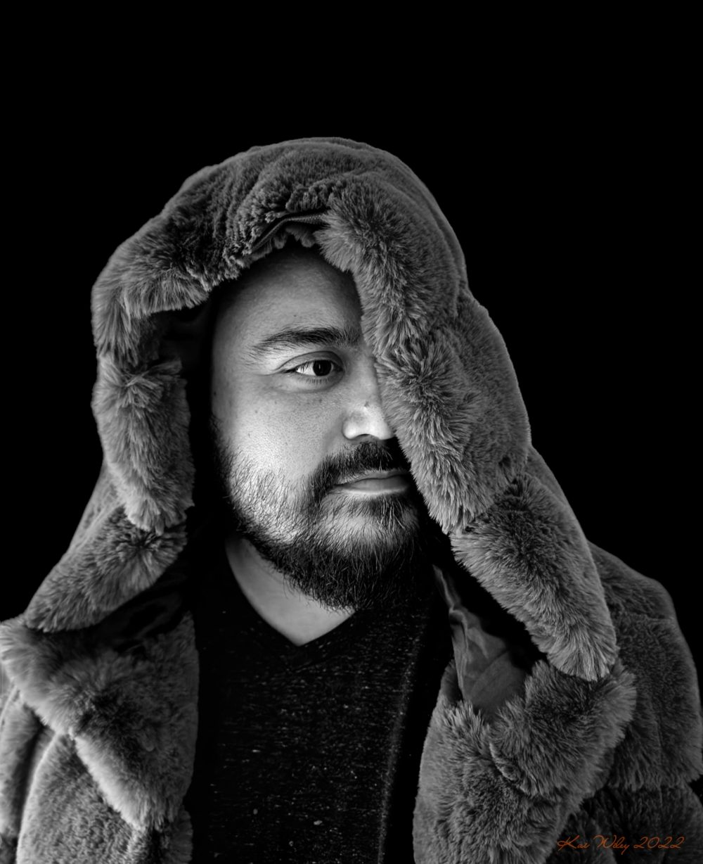 A man wearing a faux fur vest in a black and white photo with the hood slightly covering one eye.