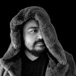 A man wearing a faux fur vest in a black and white photo with the hood slightly covering one eye.