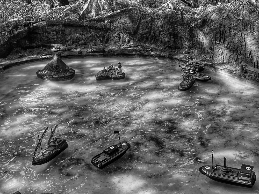 Black and white photograph of six miniature remote controlled boats scattered within a miniature water scene with a couple of isles and mountains in the background.