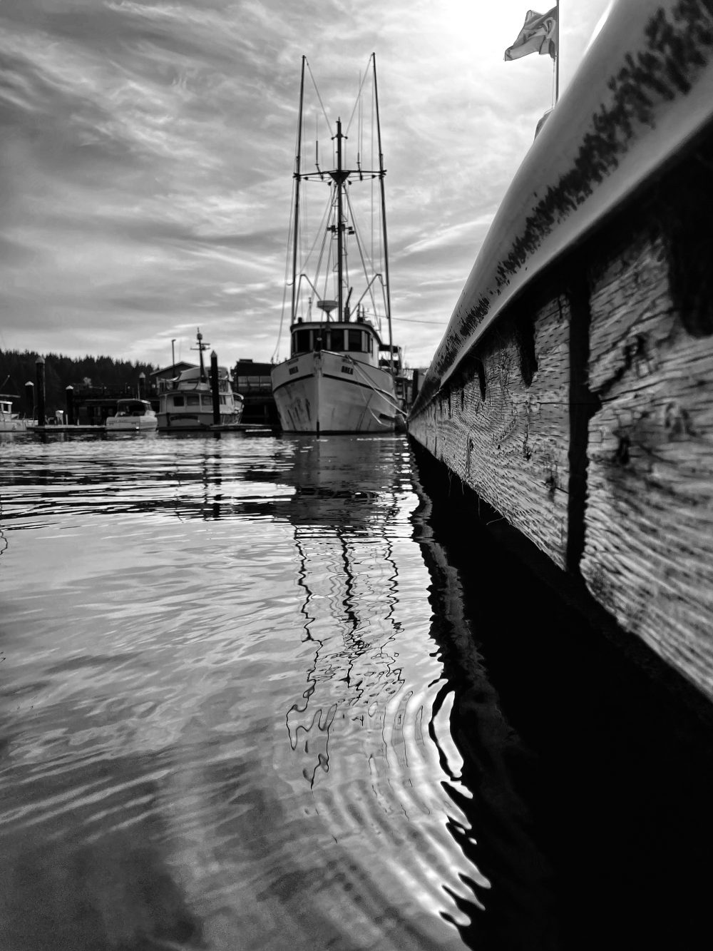 Black and white photograph of a boat docked with a flag flying in the background and ripples off the deck in the foreground.