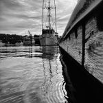 Black and white photograph of a boat docked with a flag flying in the background and ripples off the deck in the foreground.