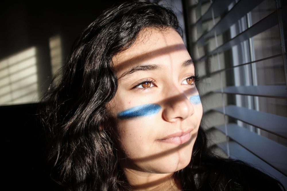 Photograph of a young woman using natural light and shadows, with blue paint under her eyes, representing war paint.
