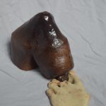 A sculpture of a burgundy water bottle that is bent down, facing the ground, being opened by part of a beige foot.