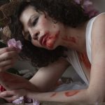 A women lying on her stomach with blood on her face, neck and arms, staring at a pink flower.