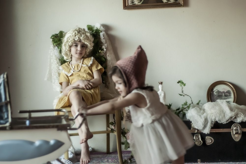 A child in a dress and a wig sitting in a chair, while a younger child walks past pushing a doll pram.