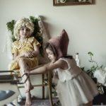 A child in a dress and a wig sitting in a chair, while a younger child walks past pushing a doll pram.