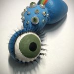 A teal bug-like creature with a large green eyeball for a head, eyelashes and multiple smaller green eyes on the body looks up past the viewer (video: process video of the construction and “making” of Apollonia”).