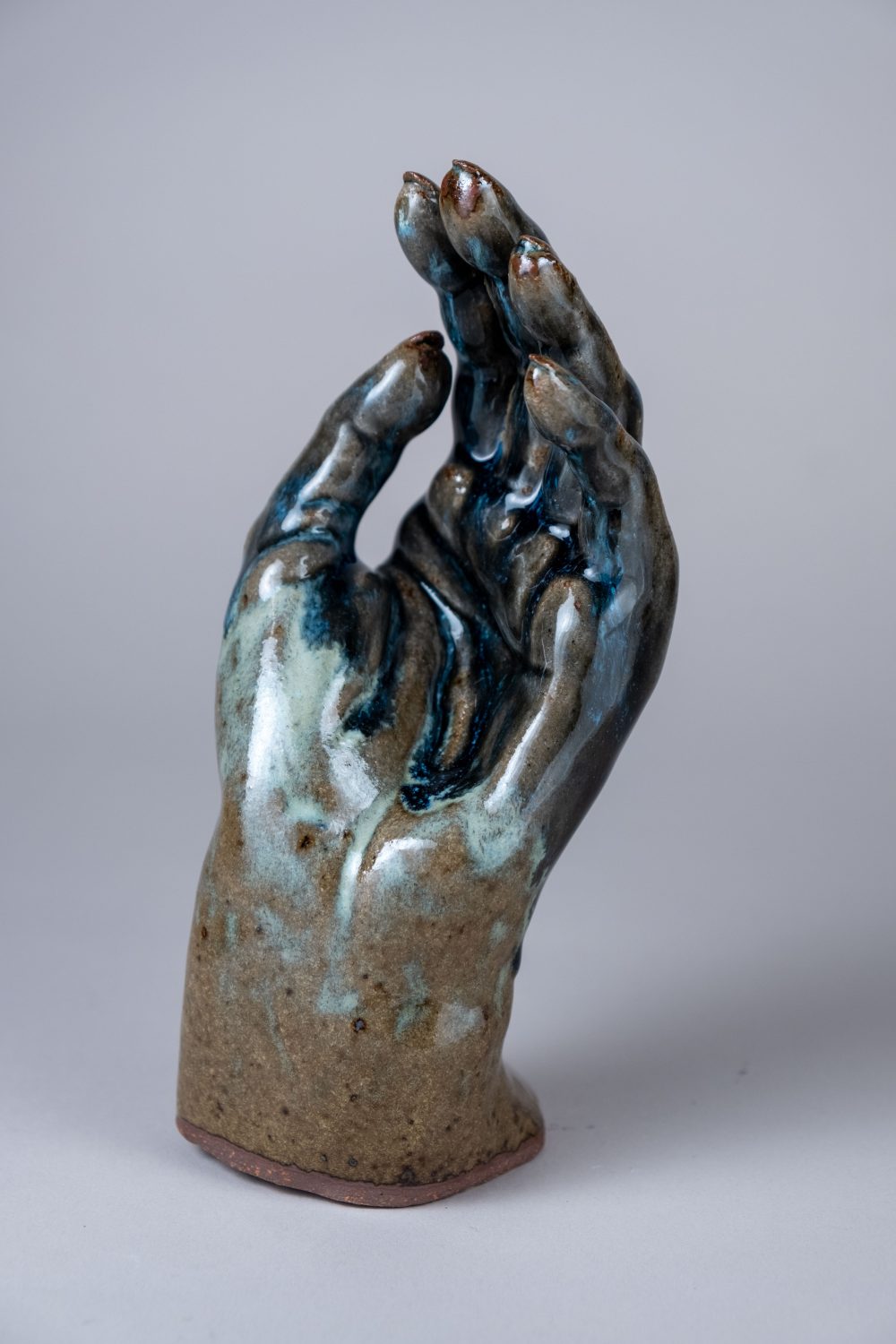 A ceramic sculpture of an upright, relaxed human hand and wrist, glazed with shades of blue and red-tinted brown.
