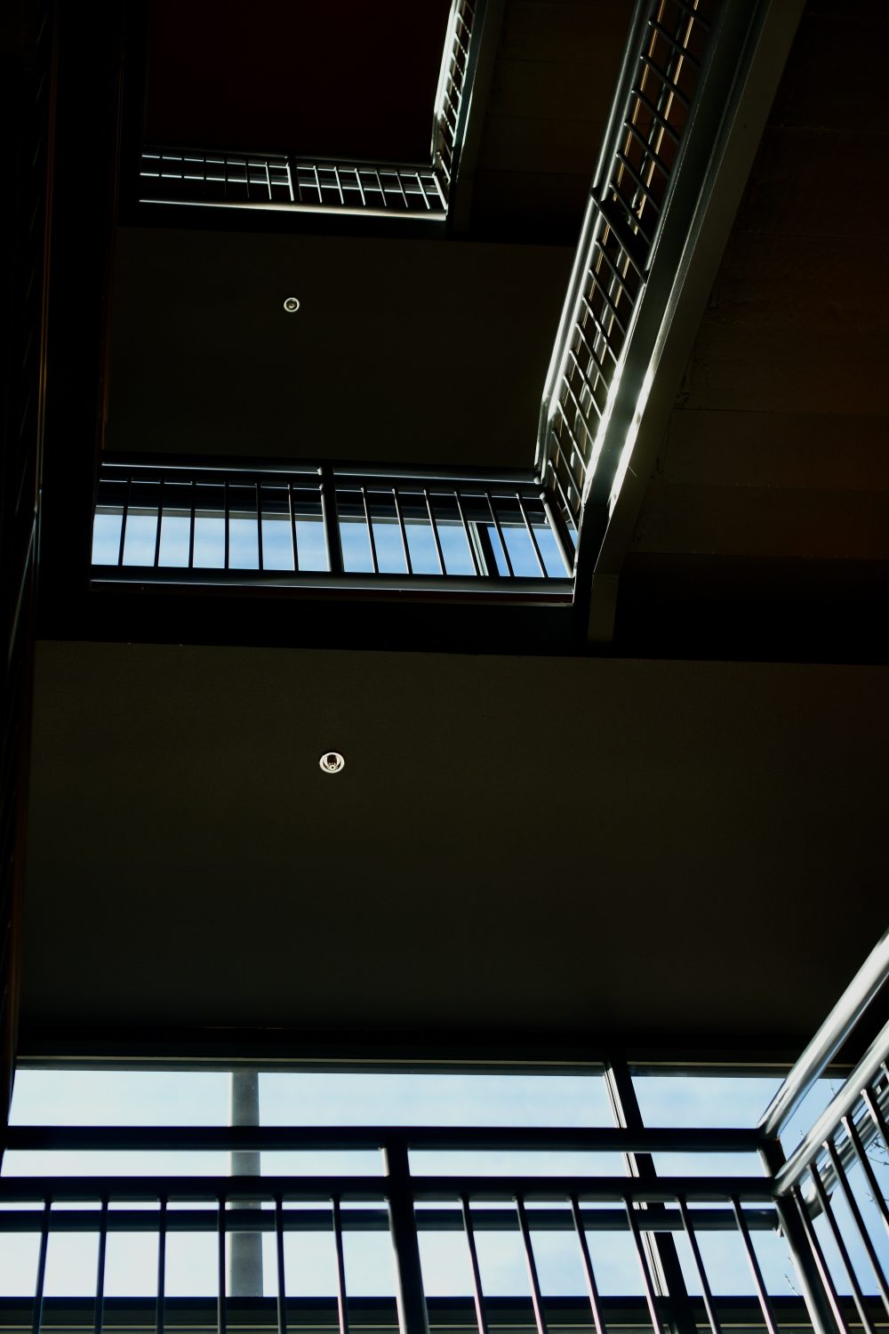 A photograph featuring light coming through a large window and shining on a staircase that goes up several flights in the inside of a building. the railing is highlighted along with a couple of recessed light fixtures on the level of each floor going up. the colors are steel gray and some white with blue sky showing through the window.