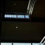 A photograph featuring light coming through a large window and shining on a staircase that goes up several flights in the inside of a building. the railing is highlighted along with a couple of recessed light fixtures on the level of each floor going up. the colors are steel gray and some white with blue sky showing through the window.