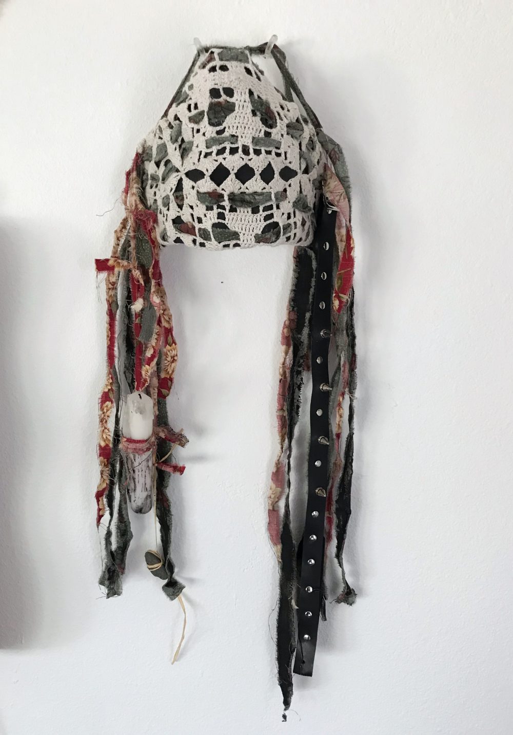 A facemask covering half the face created by a foundation of cardboard. The ground of the mask is black with a doily wrapped around the front. The holes of the doily create almost a skull-like appearance. Attached to either side of the mask are strips of torn fabric with a red floral motif. Some fabric pieces are covered in black paint. A candle is attached but not lit, dangles down with the strips of fabric. A small rock is tied with twine and attached in a similar way. A strip of leather with spikes is also attached, mimicking the strips of fabric in the piece.