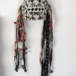 A facemask covering half the face created by a foundation of cardboard. The ground of the mask is black with a doily wrapped around the front. The holes of the doily create almost a skull-like appearance. Attached to either side of the mask are strips of torn fabric with a red floral motif. Some fabric pieces are covered in black paint. A candle is attached but not lit, dangles down with the strips of fabric. A small rock is tied with twine and attached in a similar way. A strip of leather with spikes is also attached, mimicking the strips of fabric in the piece.