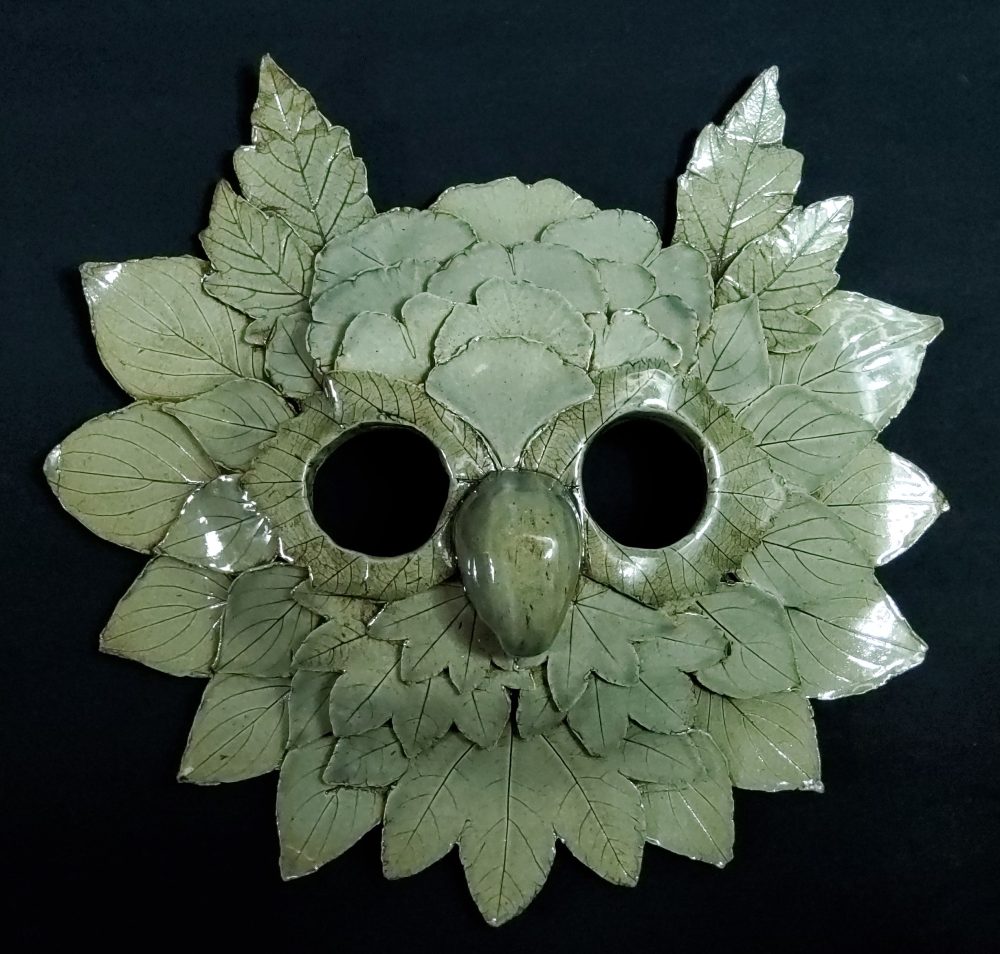 A ceramic mask with a sage green glazed, made of many clay leaves to look like an owl.