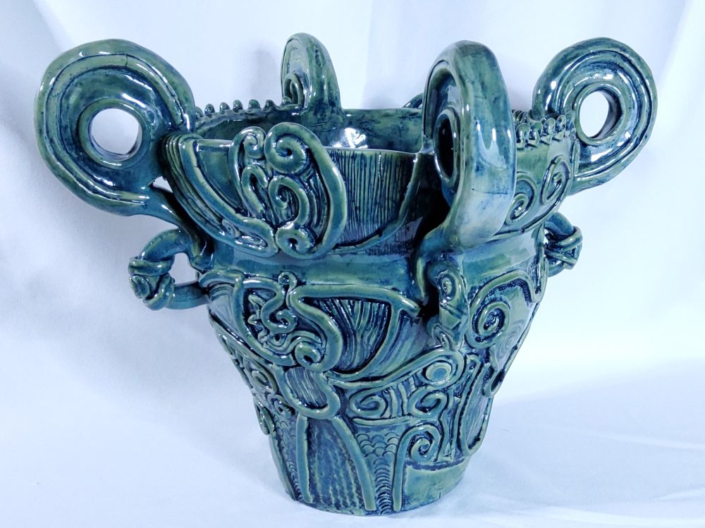 A blue green ceramic pot with four big handles and a textured pattern made of many loops and coils.