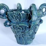 A blue green ceramic pot with four big handles and a textured pattern made of many loops and coils.