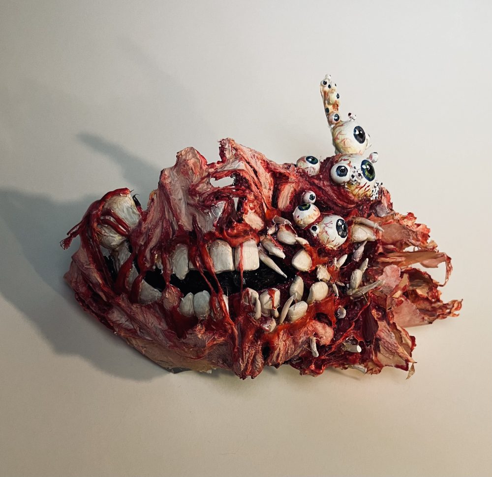 A 3D sculpture of a mask that looks like torn flesh and muscle over the jaw and teeth bones - some bones showing through the flesh with several eyes and broken bones sticking out on a white background.