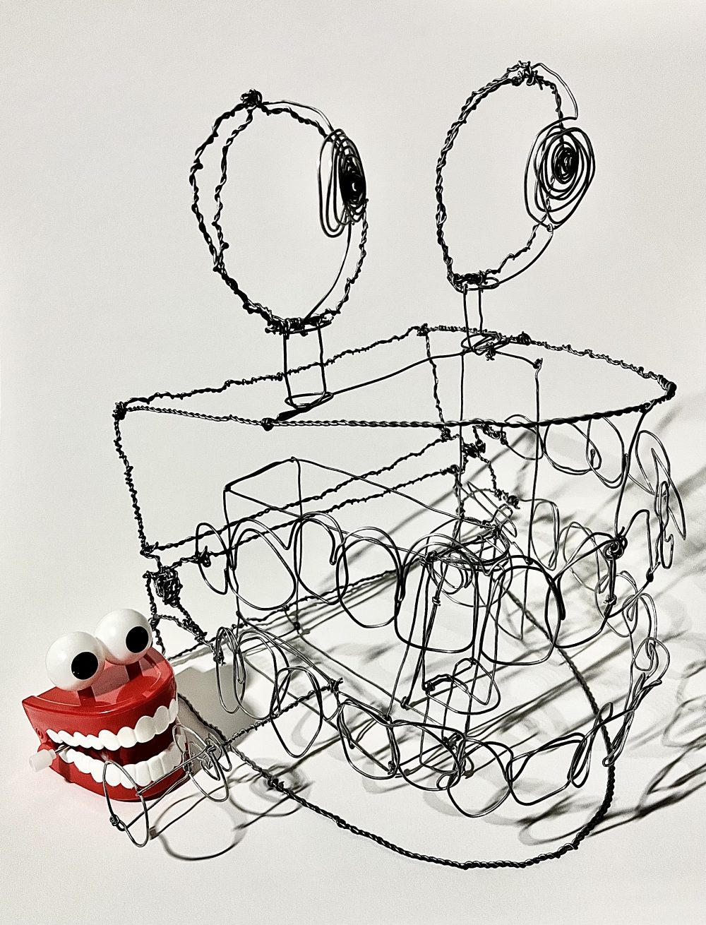 A sculpture constructed of silver wire, representing a chatter-teeth toy with large, round eyes on top with a white background.