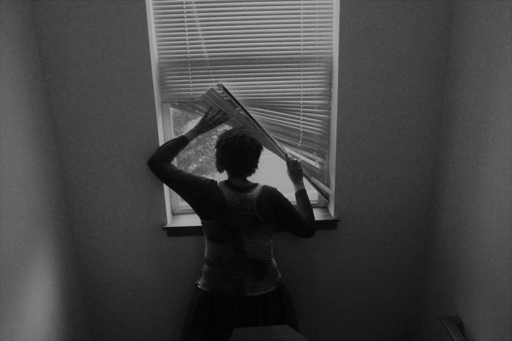 A black and white photograph showing the back of a person lifting the corner of the windowshade and looking out the window at the road and a tree.