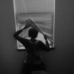 A black and white photograph showing the back of a person lifting the corner of the windowshade and looking out the window at the road and a tree.