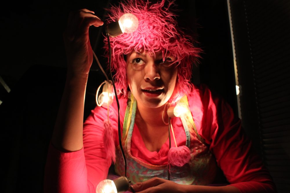Night photograph of a person dressed in a colorful outfit and monster hat with a string of bright lightbulbs draped around her neck, looking at one bulb held above her head.