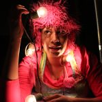 Night photograph of a person dressed in a colorful outfit and monster hat with a string of bright lightbulbs draped around her neck, looking at one bulb held above her head.