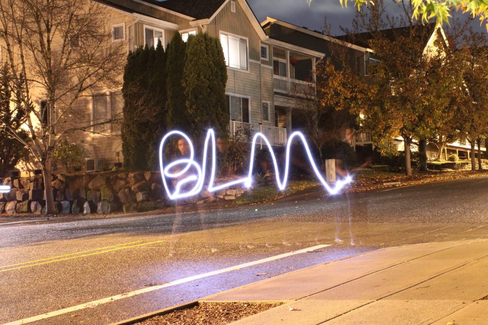 A long-exposure photograph of the transparent form of a Black woman on a residential street, using a flashlight to paint the letters BLM in the air.