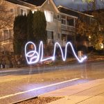 A long-exposure photograph of the transparent form of a Black woman on a residential street, using a flashlight to paint the letters BLM in the air.