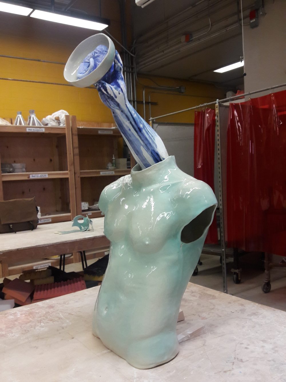 A not so small sculpture of a light blue torso with a blue and white arm coming out of the neck and holding a ceramic bowl with an ear in it.