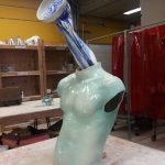 A not so small sculpture of a light blue torso with a blue and white arm coming out of the neck and holding a ceramic bowl with an ear in it.