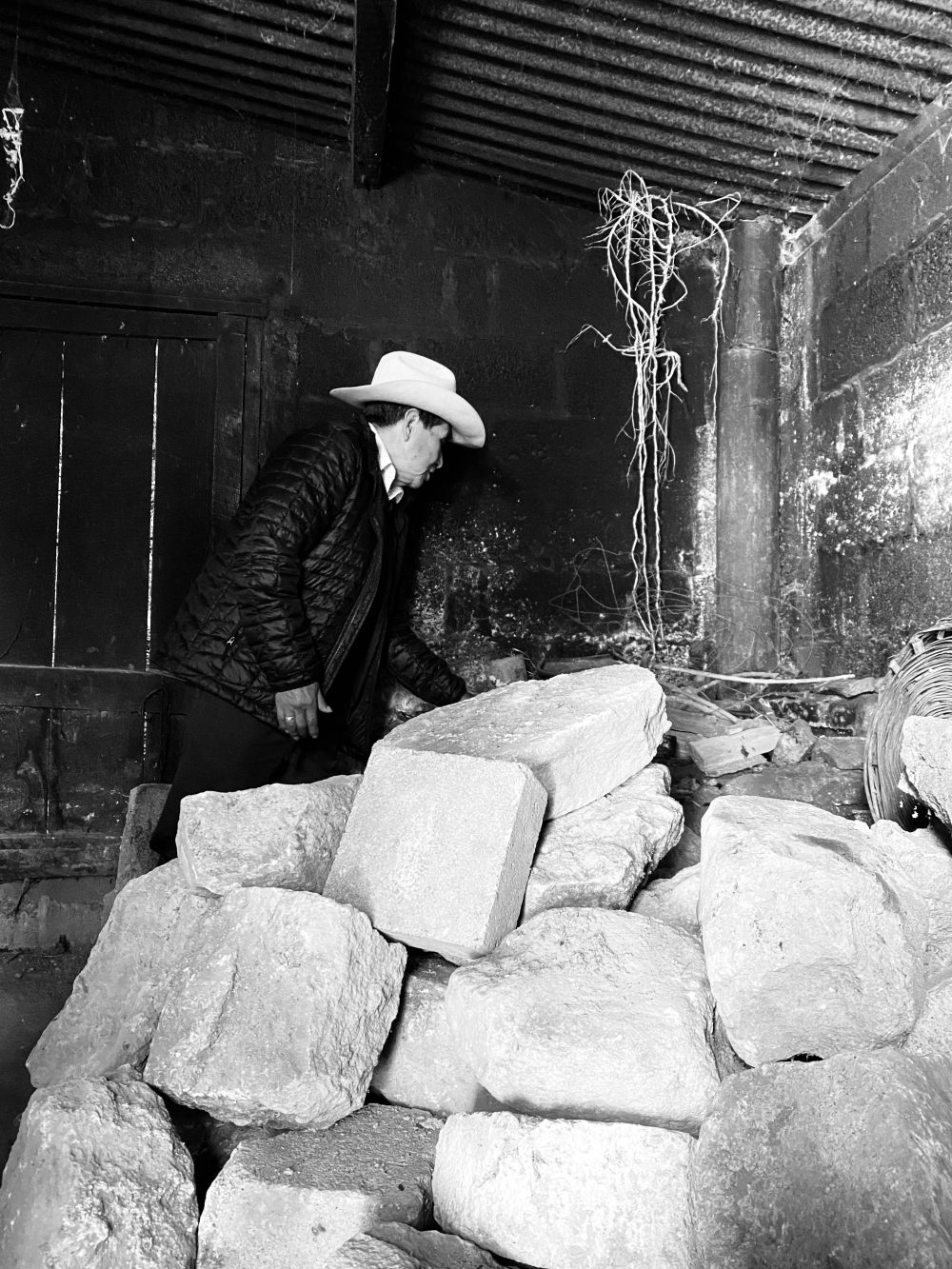 A black and white photograph with a pile of stones in the foreground and a man looking away from the camera in a black coat and white cowboy hat behind the stones.