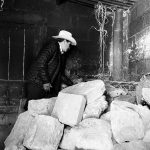 A black and white photograph with a pile of stones in the foreground and a man looking away from the camera in a black coat and white cowboy hat behind the stones.