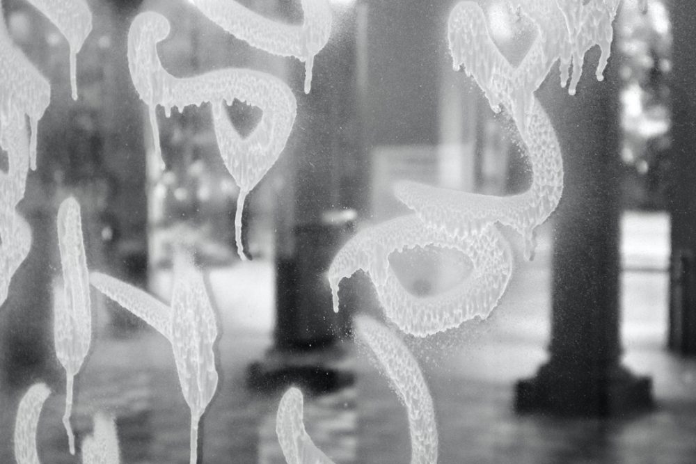 A photo in black and white of indistinct graffiti spray painted across the windows of a government building containing tall pillars and a checkered floor.