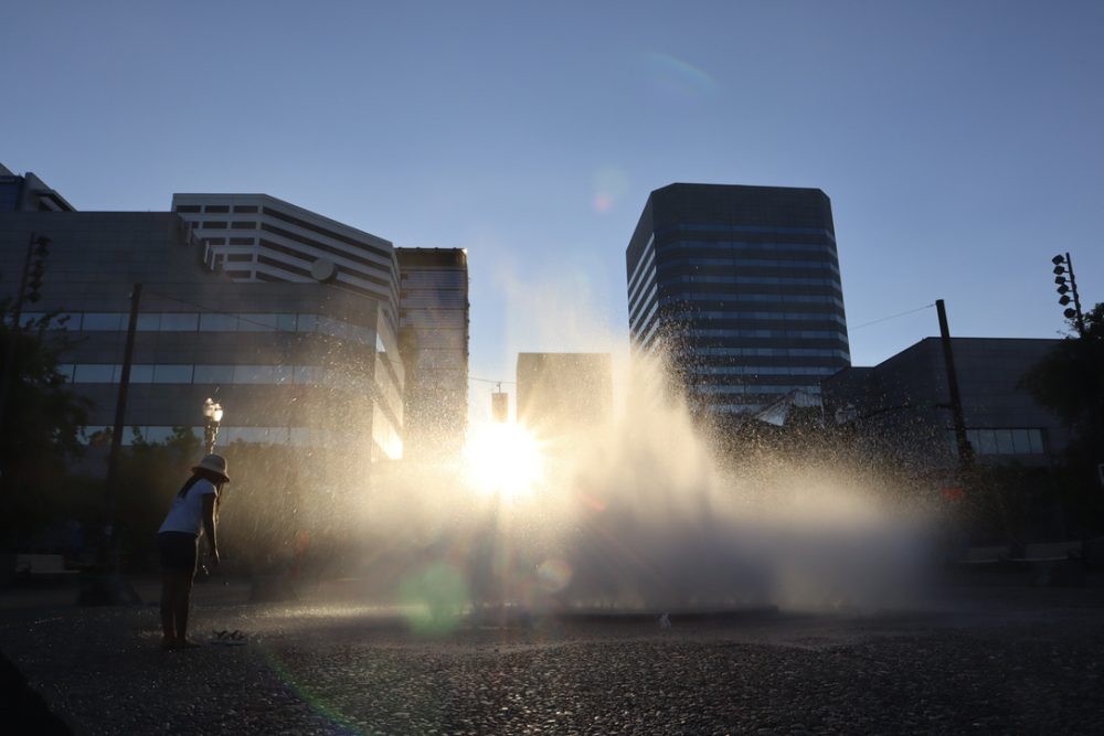 A young girl stands to the left of the Salmon Street Springs Fountain, skyscrapers tower in the background, golden light pours through the water, and a lens flare encircles the fountain.