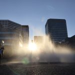 A young girl stands to the left of the Salmon Street Springs Fountain, skyscrapers tower in the background, golden light pours through the water, and a lens flare encircles the fountain.