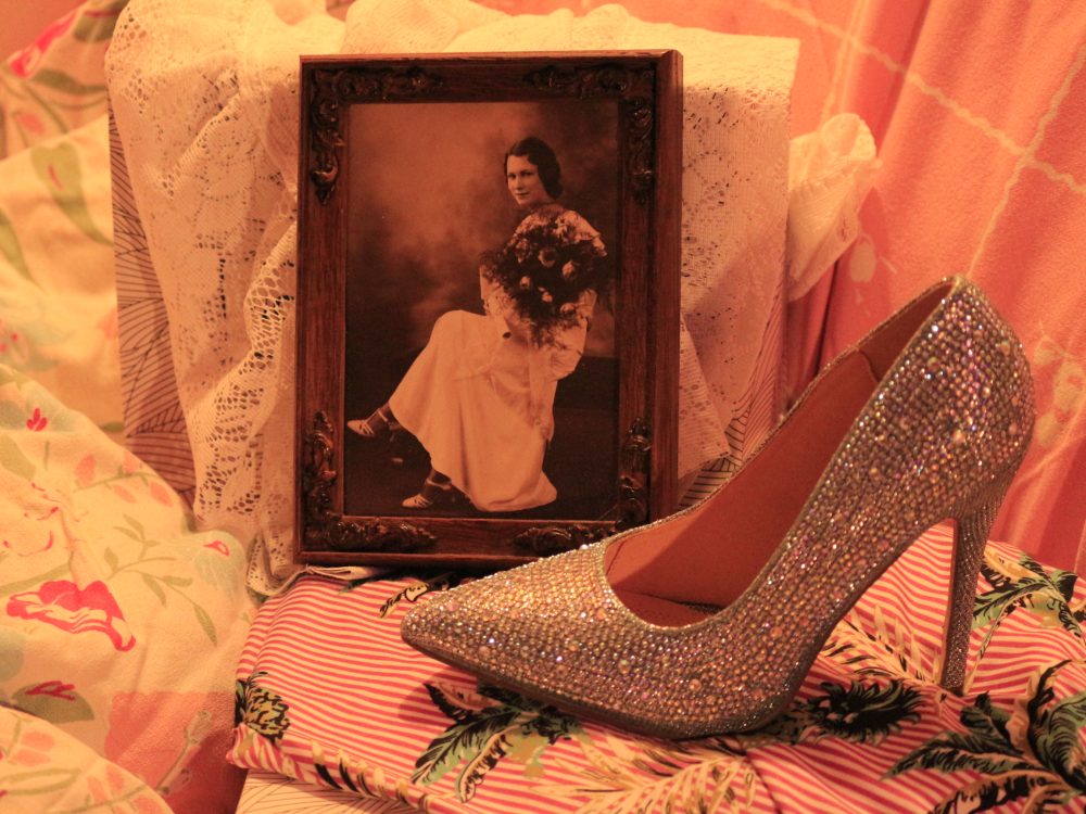 A photograph of a bejewelled high heel shoe in front of a photograph of the artist grandmother's graduation photo on a table and background of lace and floral fabrics.