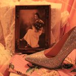 A photograph of a bejewelled high heel shoe in front of a photograph of the artist grandmother's graduation photo on a table and background of lace and floral fabrics.
