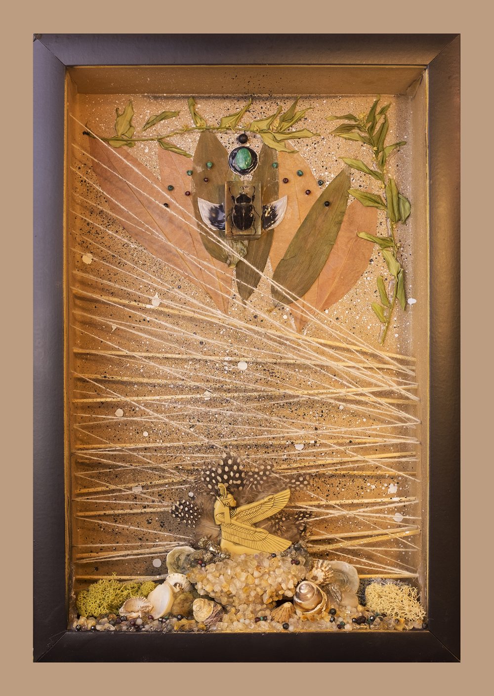 The frame is black and inside are earthen elements including semiprecious gemstones, shells, moss, an Egyptian statue at the bottom center of the box, overlapping yarn and bamboo on the inner plane, and, at the top of the sculpture, a beetle encased in resin surrounded by dried foliage.