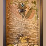 The frame is black and inside are earthen elements including semiprecious gemstones, shells, moss, an Egyptian statue at the bottom center of the box, overlapping yarn and bamboo on the inner plane, and, at the top of the sculpture, a beetle encased in resin surrounded by dried foliage.