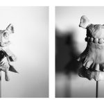 The sculpture looks like a mouse, has whiskers, long feet, trousers, a robe, and beaded necklace. It holds a jasper stone at the center of its belly.