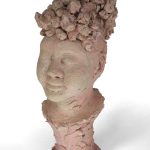 Sculpted portrait of a woman with her eyes closed into a slight smile with round cheeks, hair is pulled back into a poof of curls, details of ear piercings on both ears, the base is built of petal-like smudges pushing up into the collar and neck of the bust as slight pink stains come through in the deeper edges.